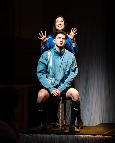 A woman with dark hair stands behind a boy displaying her hands wide on either side of his head. The boy in front discouragingly sits on a stool wearing a blue track suit.