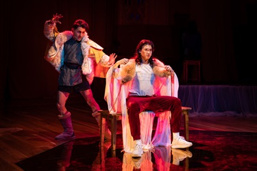 A person dressed in cultural attire stands behind and to the left of another. Their one arm is raised in the air and feet splayed in a soft side lunge. The person in front sits on a bench, removing their long robe.