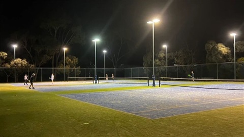 People playing tennis on grass courts at night.