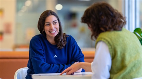 Two women sitting at a table reviewing information. One woman wears a blue jumper and smiles at the other woman