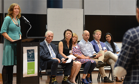 Town hall meeting with a speaker at a podium and a panel of people on chairs, listening to an audience member speak.