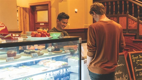 A male cafe worker looks at the register and smiles while serving a customer