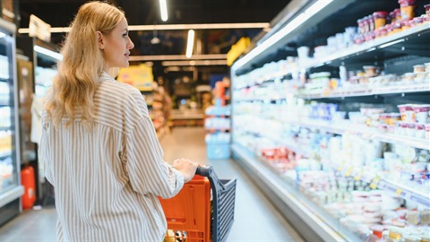 Woman with blonde hair steers a supermarket trolley and looks at food in a refrigerated cabinet with her head to the side