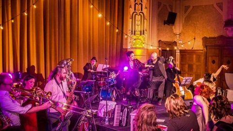 Mid-distance view of band performing on stage in indoor hall, cast in purple light and orange glow above