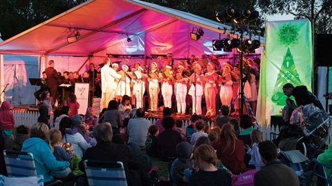 A group of people standing at microphones singing carols, underneath a large white marquee, watched by a seated gathering of familes outdoors