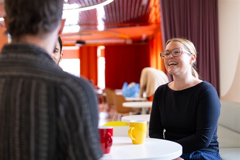 A person sitting at a cafe table with two mugs on it smiling at another person with their back towards photographer