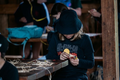 A young person wearing a black hat and tshirt that says Always Was, Always Will be is doing a weaving activity.