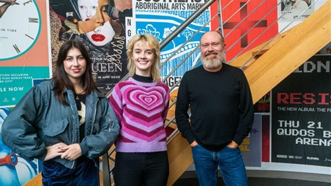 Two women and a man stand in front of large bright music posters plastered on the walls with a staircase behind them.