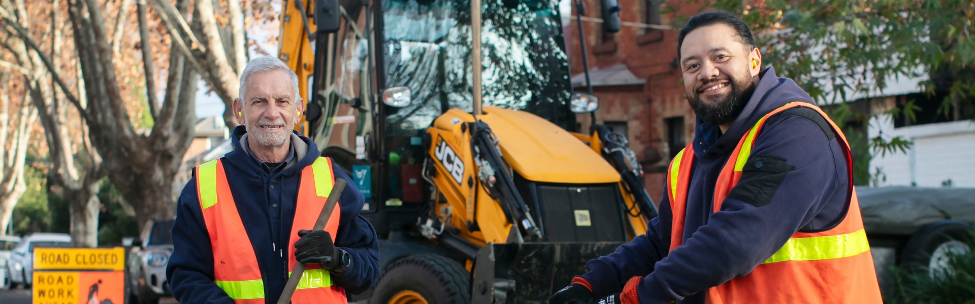 Two men wearing orange high-vis vests stand with roadwork equipment and a machine in the background
