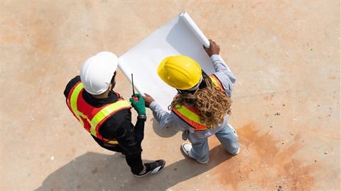 Top view of male and female engineers wearning hard hats and looking at a blueprint plan at a construction site.