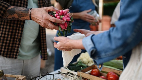 Man with tattoos on his arm holds a bunch of radishes above a cloth produce bag being held by another person