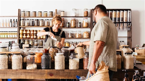 Woman staff member stands at the counter in a bulk food store and pours grain into a glass jar while a man peruses jars on the counter.