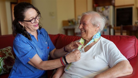 Older person wearing a white t-shirt sits next to a woman with dark hair and glasses and a blue buttoned shirt. They sit on a red couch.