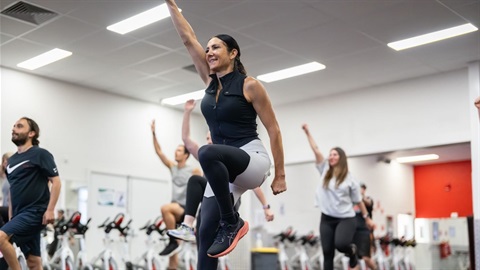 A group of men and women at a step class in a gym. They are raising their arms and knees in the air as they exercise.