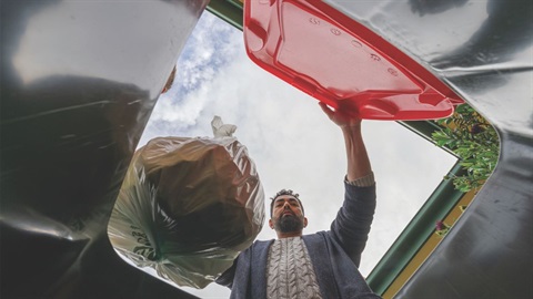 Man wearing a grey cardigan places a garbage bag full of rubbish into a waste bin with a red lid. bin
