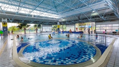 Small round pool with fencing adjoining a larger pool where parents and children stand and swim in shallow water