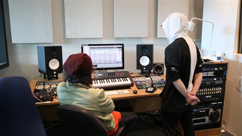 Two young people looking at a computer and sound mixing console inside a music recording studio